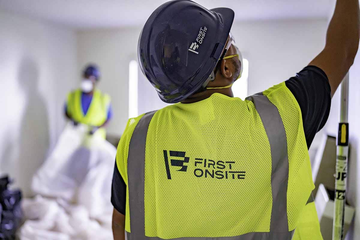 Man in First Onsite reflective vest and hard hat reaching up toward the ceiling of the room